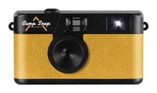 A digital camera with a black top and a textured yellow body, featuring the text "Camp Snap" on the front, a central lens, a viewfinder, and a glowing flash.
