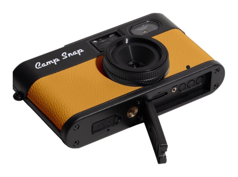 A Camp Snap digital camera with a black frame and textured yellow front panel, shown from a low angle with its bottom battery door open.