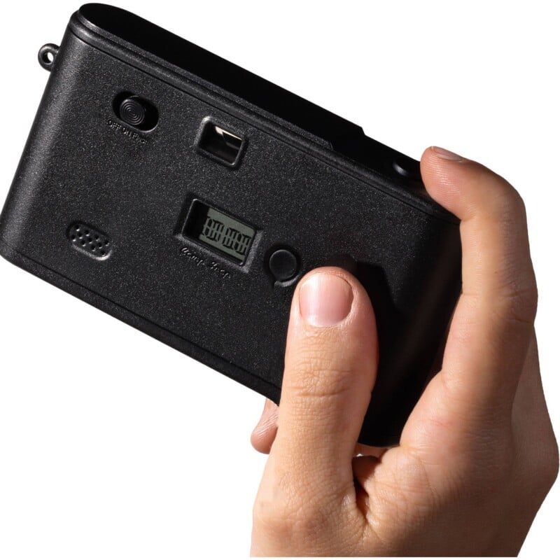 A hand holds a black, rectangular digital camera with a small LCD screen, a viewfinder, and a power switch on its back.