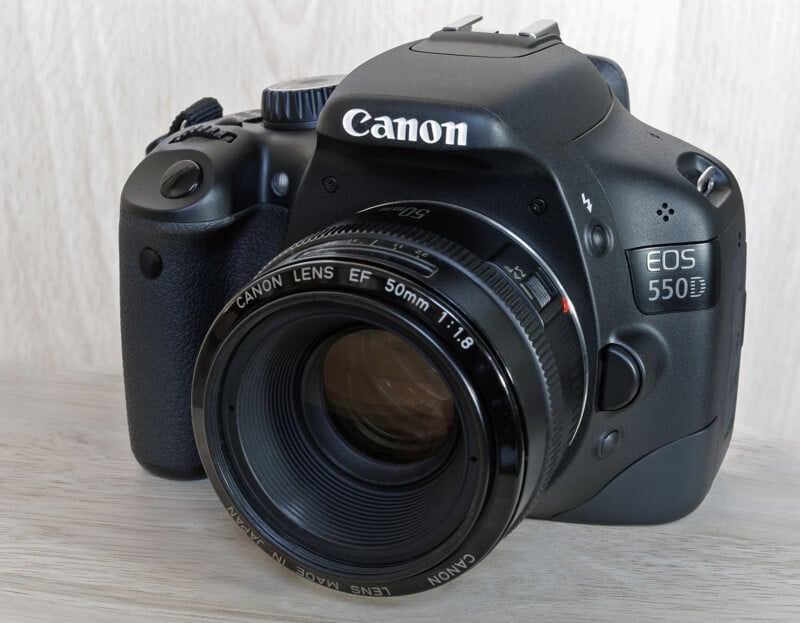 A black Canon EOS 550D DSLR camera with a 50mm f/1.8 lens attached, sitting on a light-colored wooden surface.