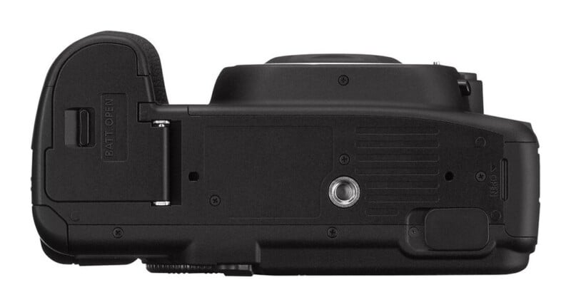 The bottom view of a black digital camera, showing the battery compartment door, a central tripod mount, and a small port cover.