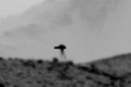 A blurry, dark, tripod-like silhouette stands atop a rocky, uneven ridge against a pale, overcast sky.