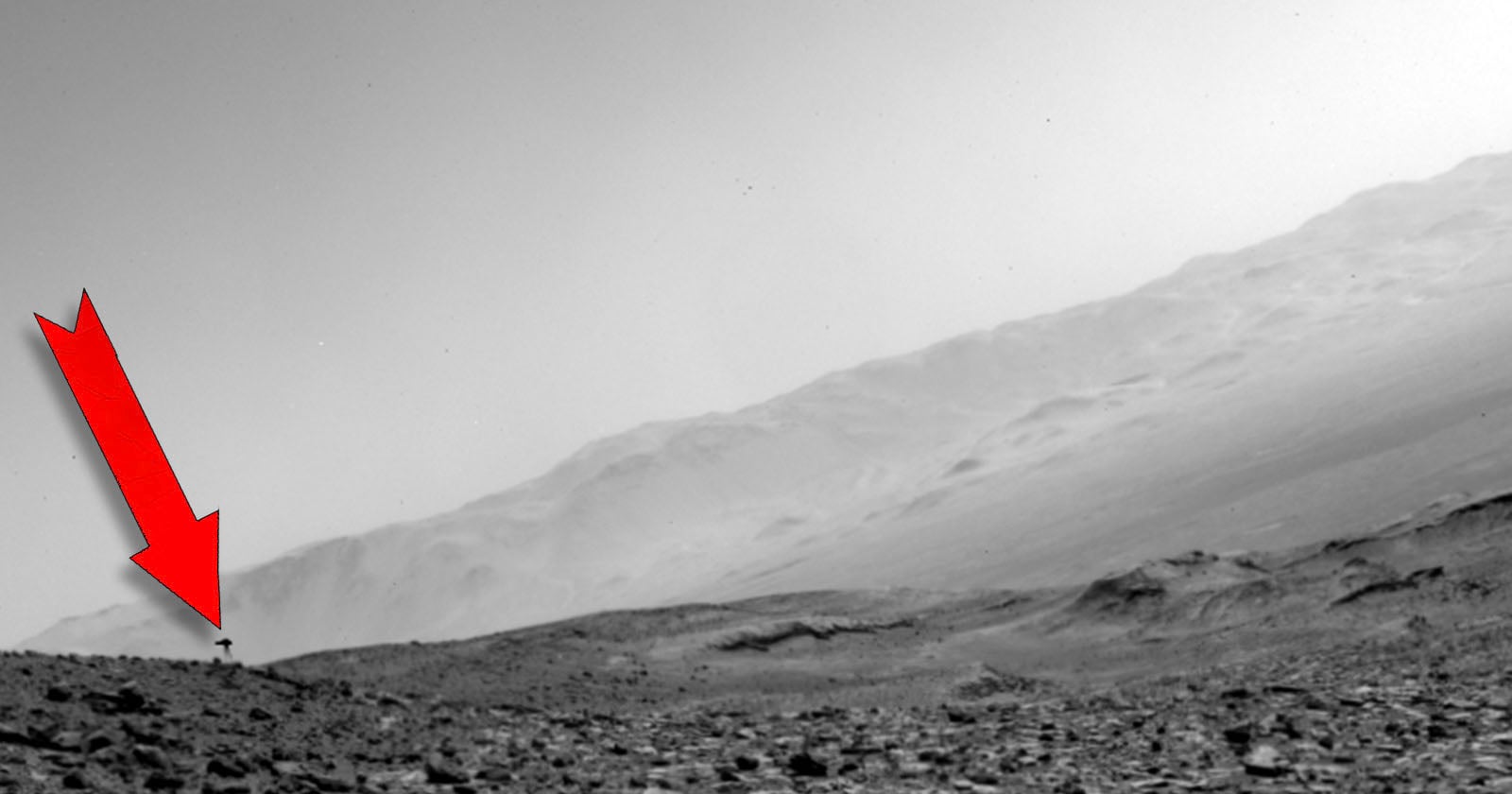 A red arrow points to a small, dark, T-shaped object resting on a rocky, barren landscape beneath a large, sloping hill.