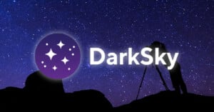 A silhouette of a person using a camera on a tripod stands on a rocky hill against a starry night sky, with a DarkSky logo.