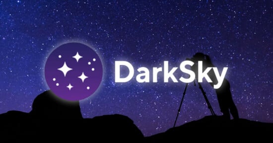 A silhouette of a person using a camera on a tripod stands on a rocky hill against a starry night sky, with a DarkSky logo.