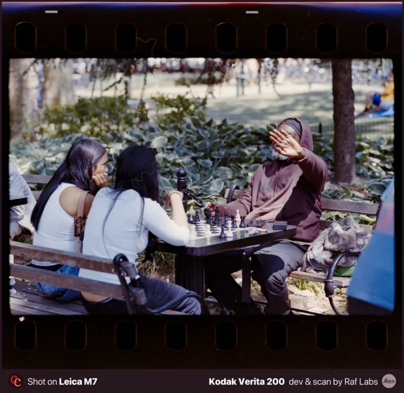 Two women sit at a park bench playing chess against a man who is holding his hand up to his face.