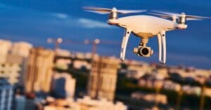 A white quadcopter drone with a camera gimbal flies in front of a blurred city skyline at dusk.