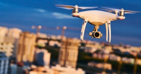 A white quadcopter drone with a camera gimbal flies in front of a blurred city skyline at dusk.