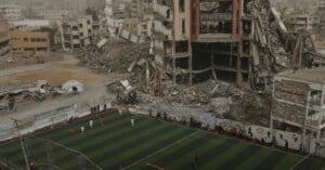 A soccer match takes place on a small turf field surrounded by heavily damaged and collapsed buildings.