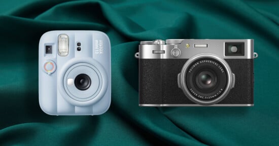A light blue Instax mini 13 instant camera and a silver and black Fujifilm X100V digital camera sit side-by-side on a dark green fabric background.