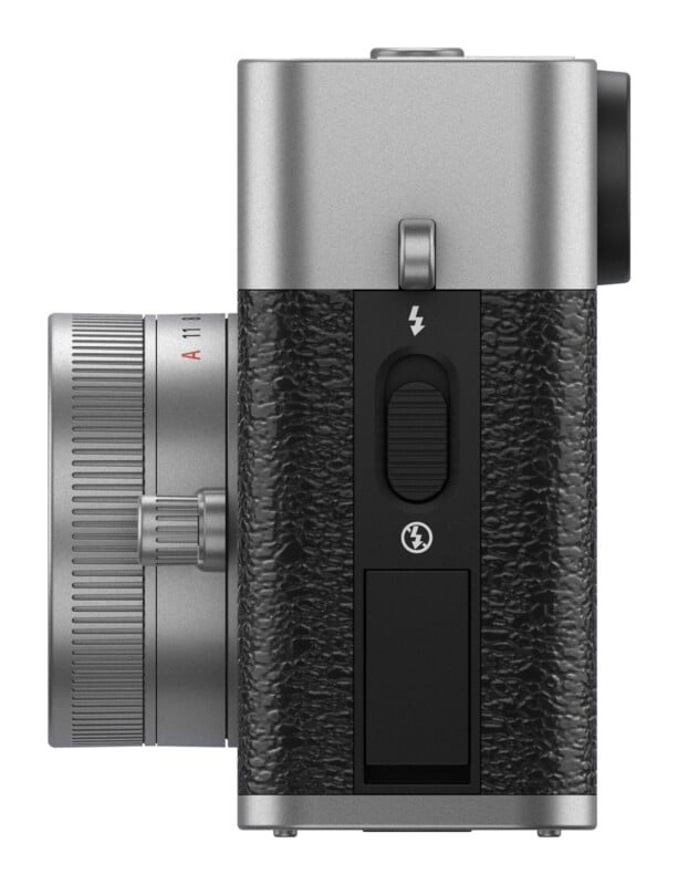 A side view of a silver and black camera body with a lens attached, showing a flash release switch and a port cover on the textured grip.