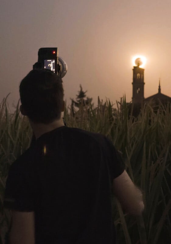 A person in a black shirt stands in a field of tall grass, holding a camera up to photograph the sun setting behind a church tower.