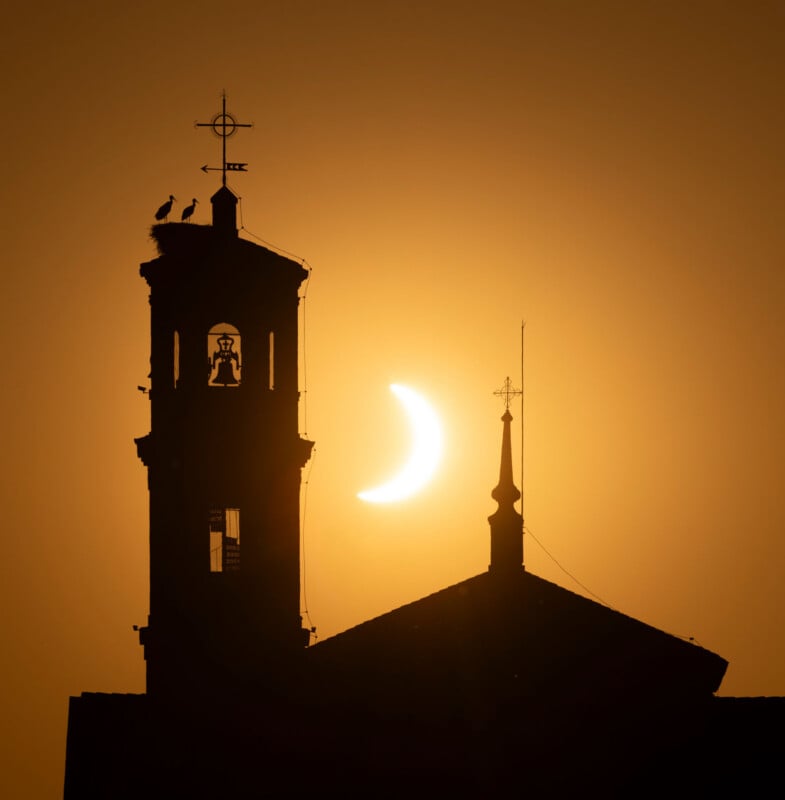 A partial solar eclipse appears behind the silhouette of a church bell tower with a stork nest on top and a smaller adjacent spire.