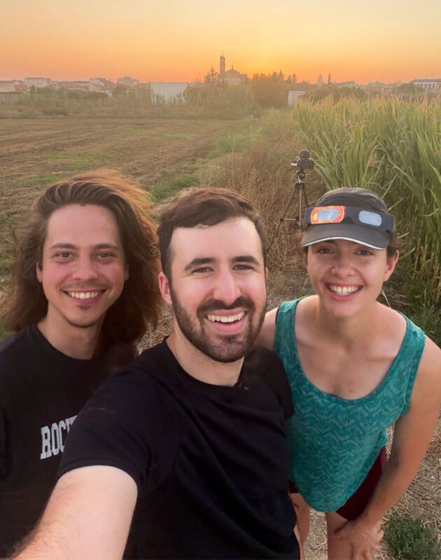 Three people smile for a selfie in a field at sunset, with a camera on a tripod visible in the background.