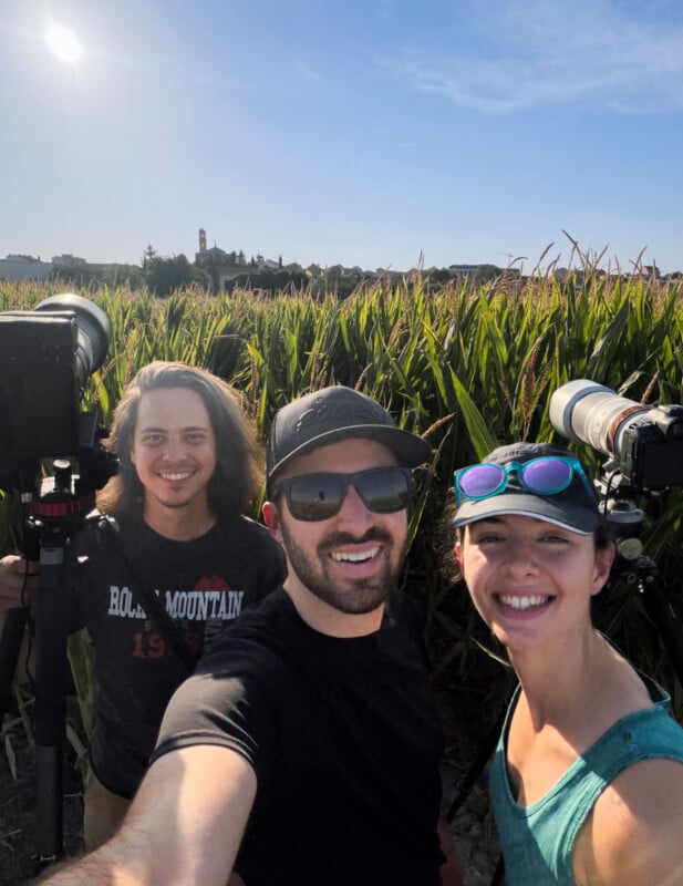 Three people smile for a selfie in a cornfield with two cameras on tripods visible in the background.