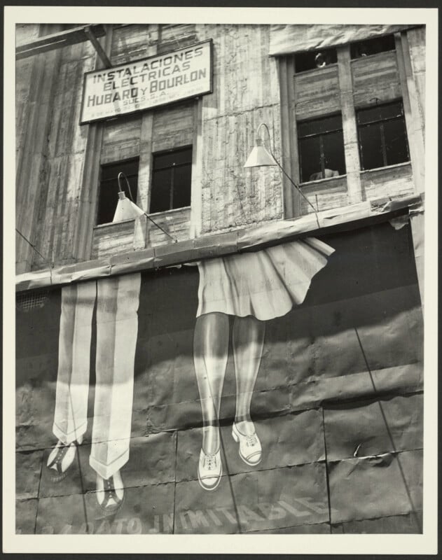 A black-and-white photograph shows a building facade with a sign for "Instalaciones Eléctricas Hubard y Bourlon" above a large advertisement featuring painted legs and feet wearing shoes.