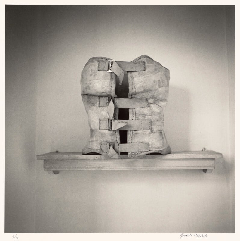 A black-and-white photograph shows a worn, vintage orthopedic corset with four buckled straps resting on a simple wooden shelf.