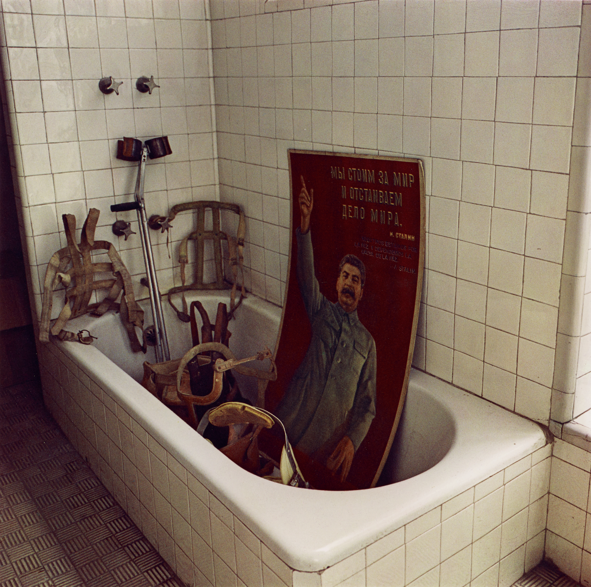 A bathtub filled with orthopedic harnesses, crutches, and a poster of Joseph Stalin pointing upward.