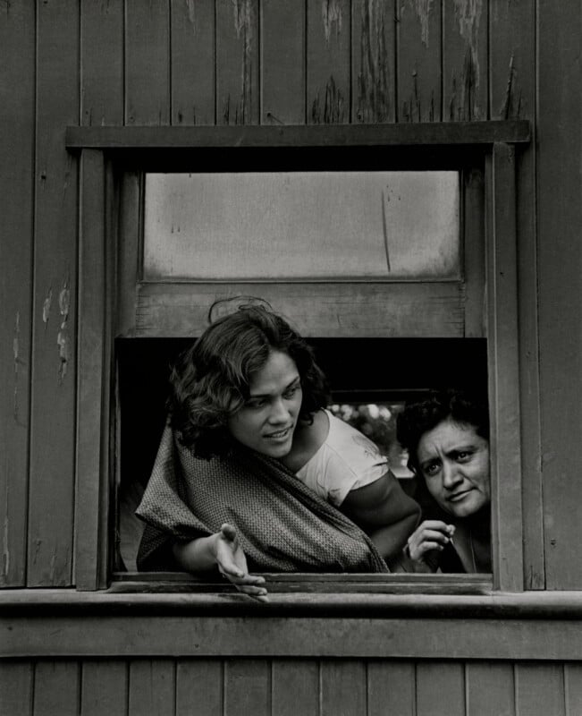 Two women look out from a rectangular window in a wooden wall, with the woman on the left gesturing with her hand.