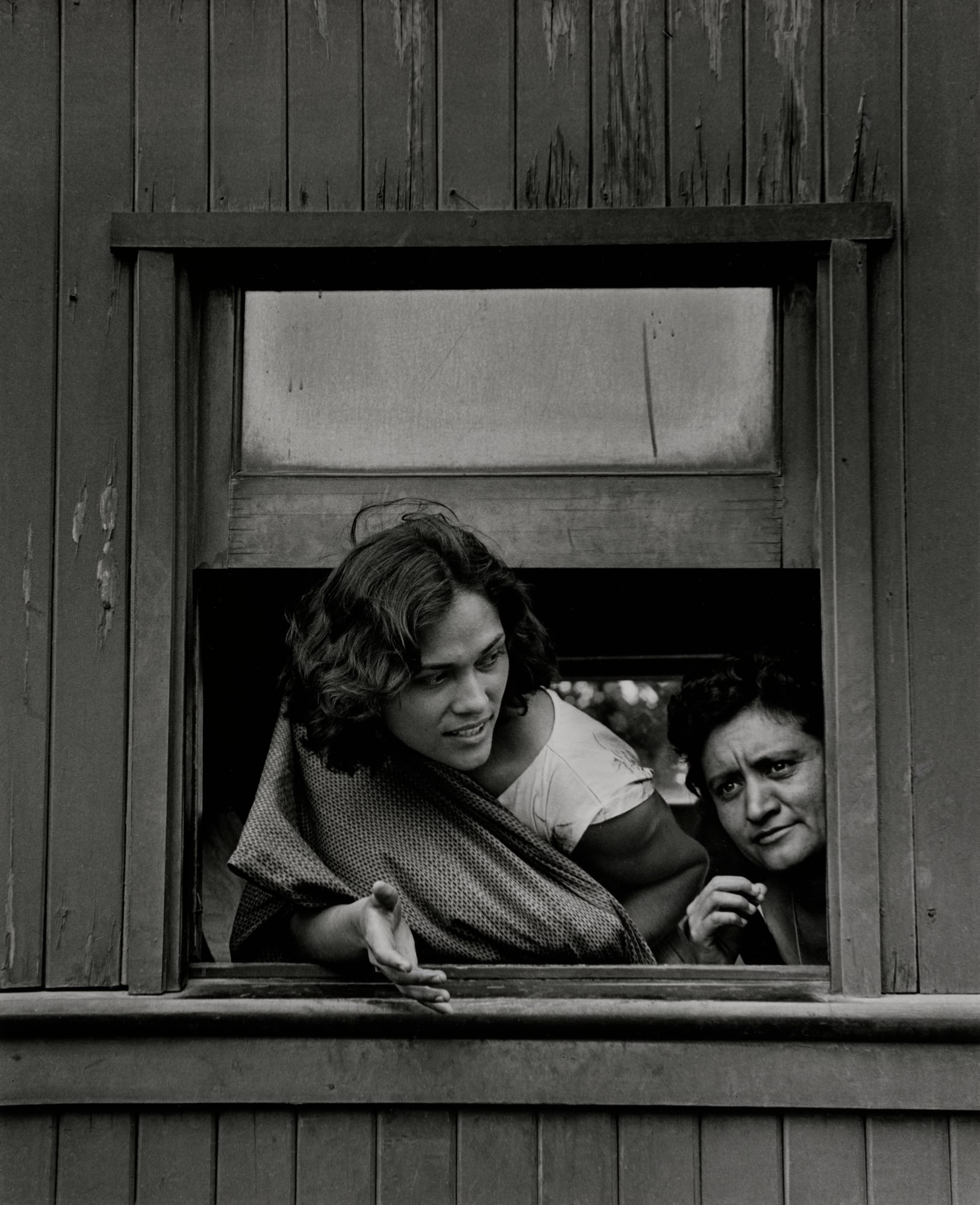 Two women look out from a rectangular window in a wooden wall, with the woman on the left gesturing with her hand.