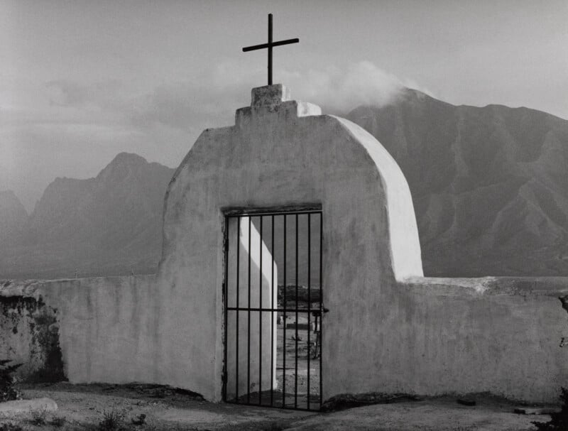 A black-and-white shot of a white stucco arched gate topped with a cross, set against a backdrop of hazy mountains.