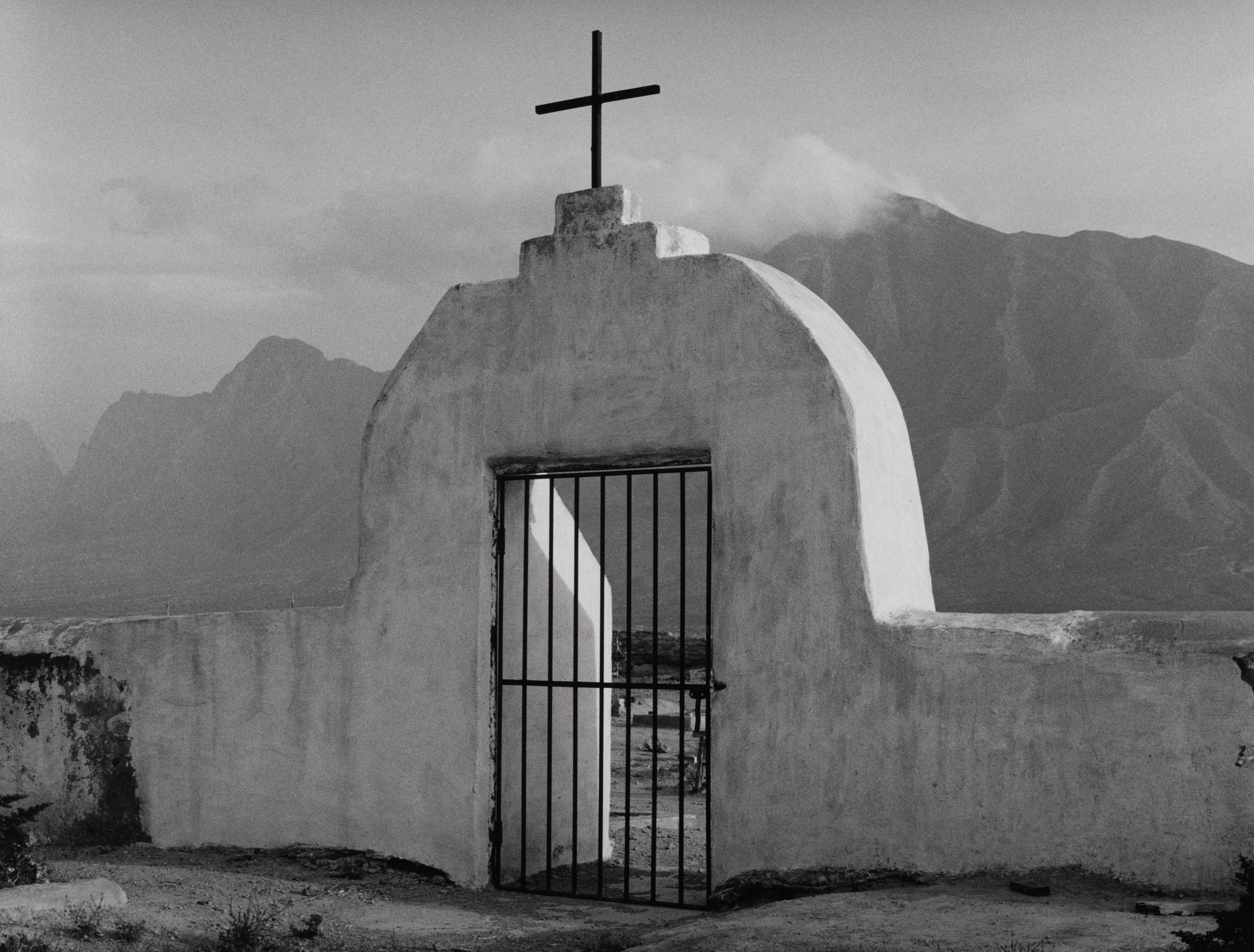 A black-and-white shot of a white stucco arched gate topped with a cross, set against a backdrop of hazy mountains.