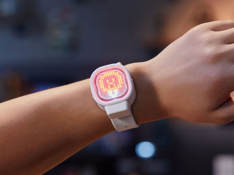 A person's arm wearing a white digital watch with a square, glowing red LED display showing a pixelated face.