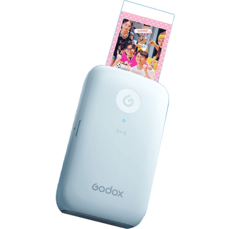 A light blue portable photo printer with a Godox logo, printing a small photograph featuring a group of people celebrating a birthday.