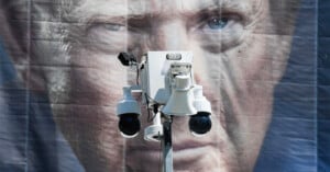 A white security camera unit with two dome cameras and a speaker is positioned in front of a large, blurry banner featuring a close-up portrait of a man.
