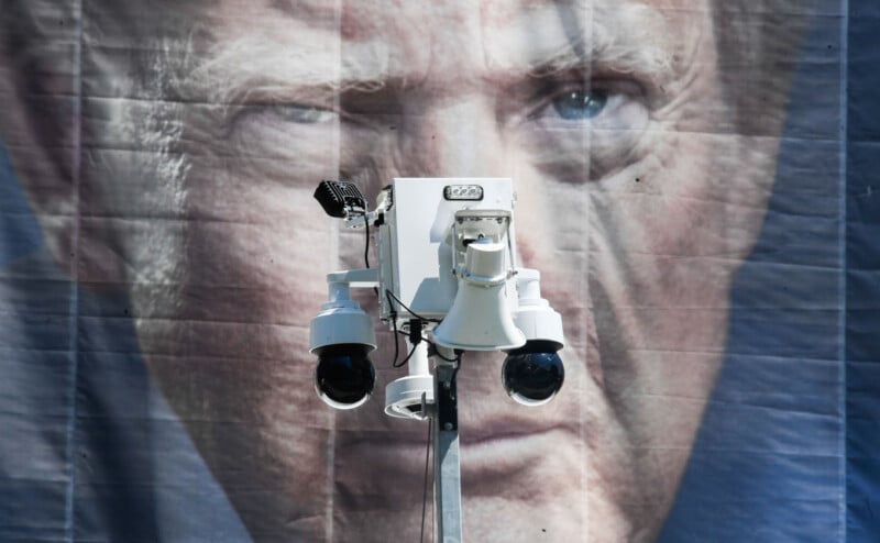 A security camera unit with two dome cameras and a speaker is mounted on a pole in front of a large banner featuring a close-up portrait of Donald Trump.