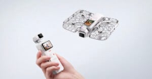 A hand holds a white handheld gimbal camera with a screen, while a matching white drone with a protective cage flies in the air nearby.