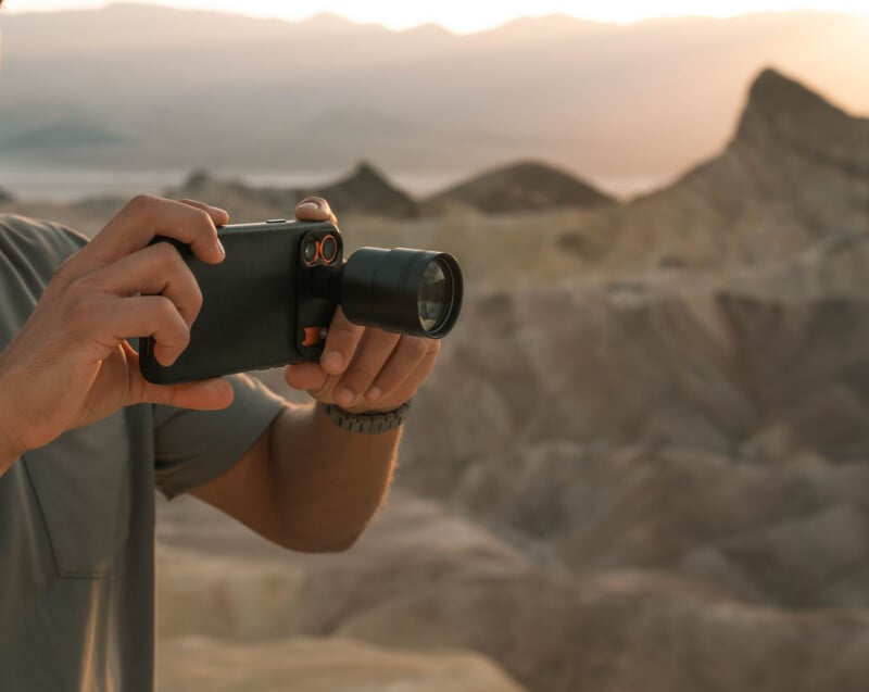 A person holds a smartphone equipped with a large external lens attachment while photographing a desert landscape at sunset.