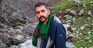 A man with a dark beard and short hair wears a green patterned scarf and a dark blue jacket, standing outdoors in a rocky, natural landscape.