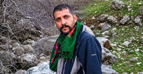 A man with a dark beard and short hair wears a green patterned scarf and a dark blue jacket, standing outdoors in a rocky, natural landscape.