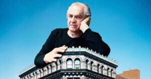 A man with gray hair rests his chin on his hand while leaning over the roof of a large, ornate stone building against a blue sky.
