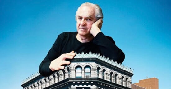 A man with gray hair rests his chin on his hand while leaning over the roof of a large, ornate stone building against a blue sky.