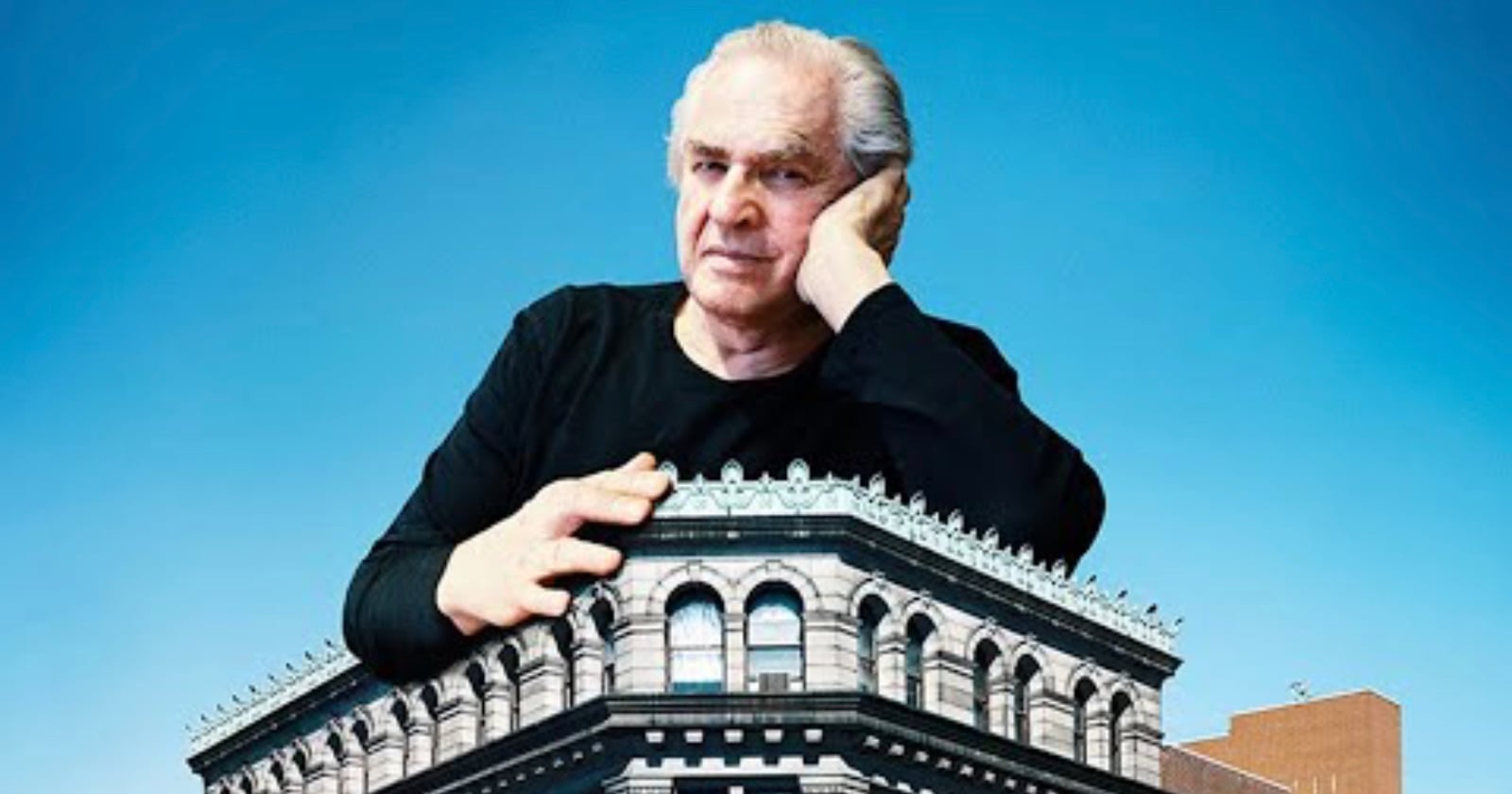 A man with gray hair rests his chin on his hand while leaning over the roof of a large, ornate stone building against a blue sky.