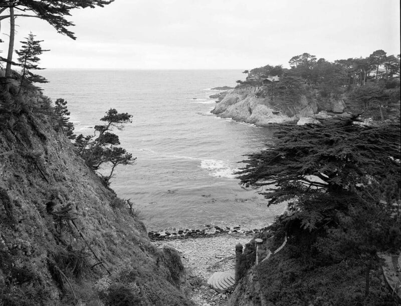 A rocky cove with a pebble beach and stone stairs leads to the ocean, framed by steep cliffs and cypress trees.