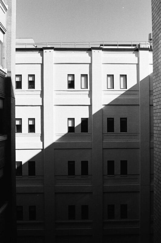 A black and white view of a multi-story building facade with symmetrical windows, partially obscured by a sharp diagonal shadow.