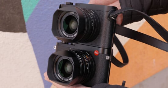 A person holds two black Leica cameras stacked vertically against a colorful painted wall.