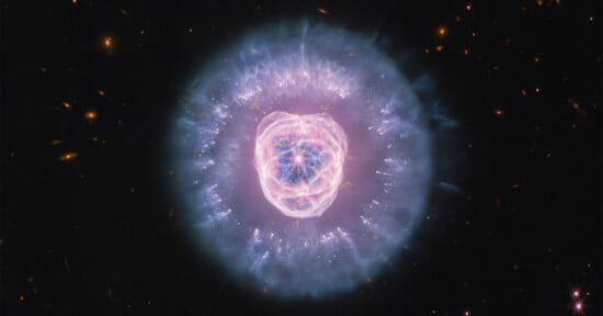 The Eskimo Nebula glows in space with a bright, pink-tinted inner core surrounded by a large, translucent blue shell of gas.