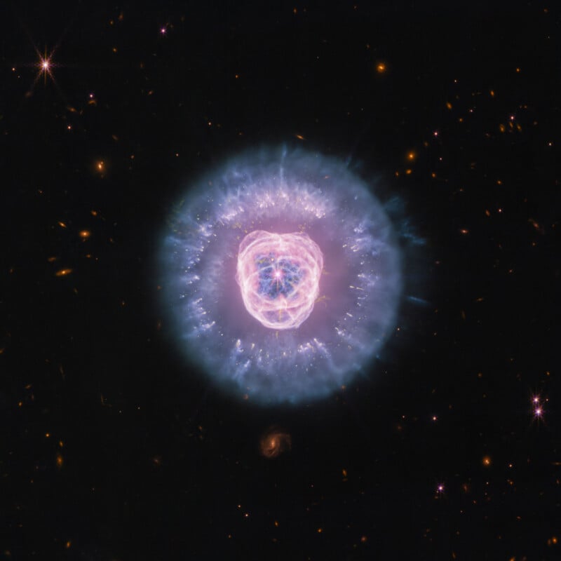 A glowing, spherical nebula with a bright, pink, heart-shaped inner structure sits centered against a dark background of distant stars and galaxies.