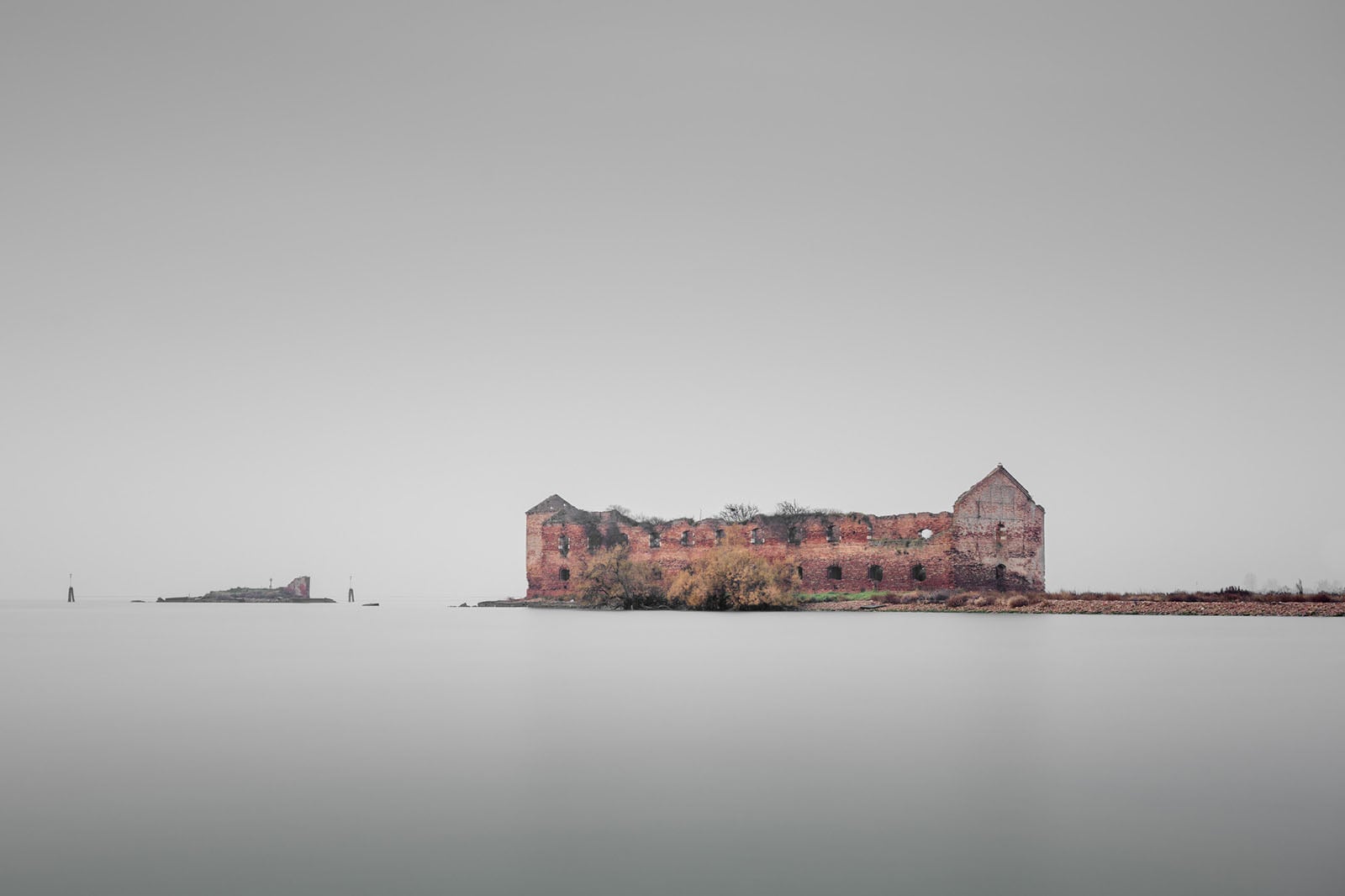 A long, ruined brick building sits on a flat, desolate island surrounded by calm, grey water under a hazy, overcast sky.