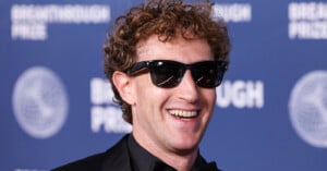 Mark Zuckerberg with curly hair and sunglasses smiles while wearing a black suit against a blue Breakthrough Prize backdrop.
