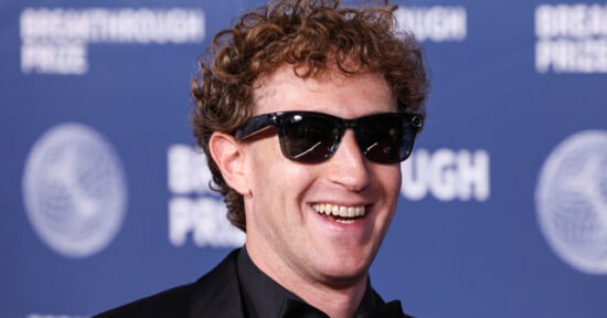 Mark Zuckerberg with curly hair and sunglasses smiles while wearing a black suit against a blue Breakthrough Prize backdrop.