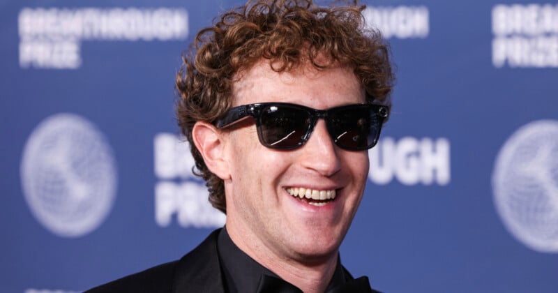 Mark Zuckerberg with curly hair and sunglasses smiles while wearing a black suit against a blue Breakthrough Prize backdrop.
