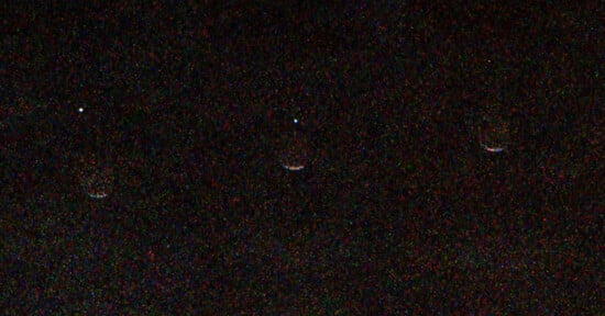 Three bright, white points of light are arranged horizontally against a dark, grainy background, each with a faint, colorful, semi-circular arc beneath it.
