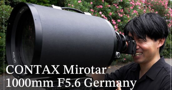 A person smiles while looking through a camera attached to a very large, black, cylindrical Carl Zeiss Mirotar 1000mm F5.6 lens.