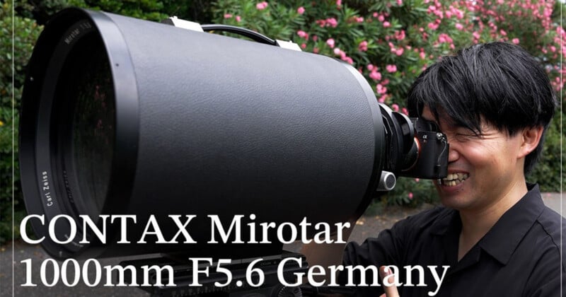A person smiles while looking through a camera attached to a very large, black, cylindrical Carl Zeiss Mirotar 1000mm F5.6 lens.