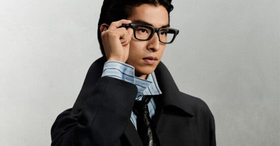 A young man with dark hair wears black-rimmed glasses, a light blue striped shirt, a dark tie, and a dark coat, with his hand resting on the frame of his glasses.